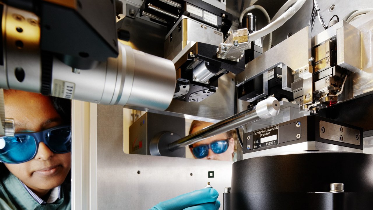 Wissenschaftlerin mit Schutzbrille greift mit einem kleinen Utensil in der Hand in eine Apparatur
